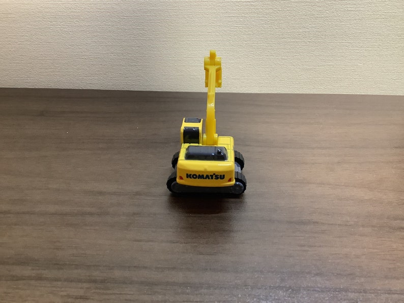 May include: A yellow Komatsu toy excavator with black tracks and a movable arm. The toy has the Komatsu logo in black on the front. The toy is on a brown wooden surface.