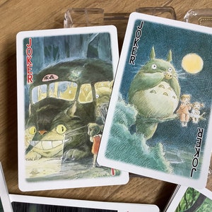 May include: Two "Joker" playing cards featuring illustrations of characters from the animated film "My Neighbor Totoro." One card shows Totoro as a bus, the other shows Totoro with children. The cards are on a wooden surface.
