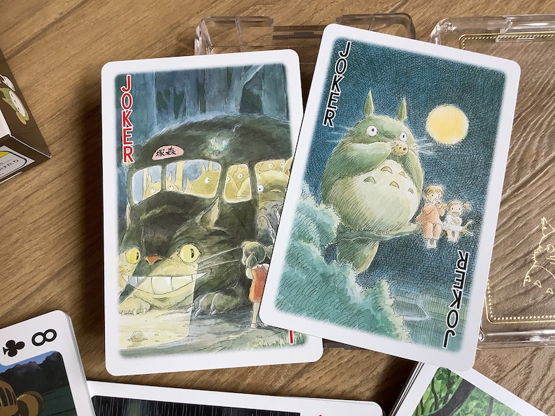 May include: Two "Joker" playing cards featuring illustrations of characters from the animated film "My Neighbor Totoro." One card shows Totoro as a bus, the other shows Totoro with children. The cards are on a wooden surface.