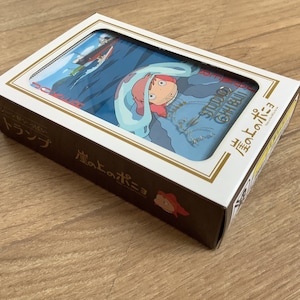May include: A rectangular box of playing cards featuring Ponyo on the cover. The box is white and dark brown with Japanese text and a graphic illustration of a character with pink hair and a blue cloak. The Studio Ghibli logo is visible.