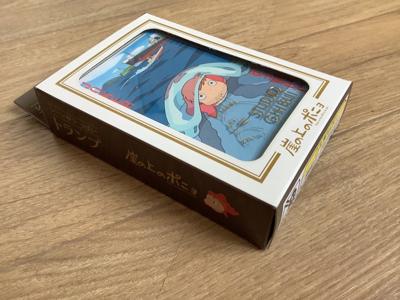 May include: A rectangular box of playing cards featuring Ponyo on the cover. The box is white and dark brown with Japanese text and a graphic illustration of a character with pink hair and a blue cloak. The Studio Ghibli logo is visible.