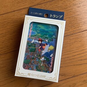 May include: A deck of playing cards featuring a colorful illustration of a character flying on a broomstick over a town. The card box has Japanese text and a white frame with gold accents. The top of the box has a blue tag with Japanese text.