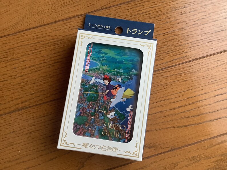 May include: A deck of playing cards featuring a colorful illustration of a character flying on a broomstick over a town. The card box has Japanese text and a white frame with gold accents. The top of the box has a blue tag with Japanese text.