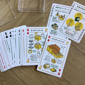 May include: A deck of playing cards with a clear plastic case. The cards feature traditional suits and numbers, along with illustrated cards with Japanese text and diagrams. The cards are on a wooden surface.