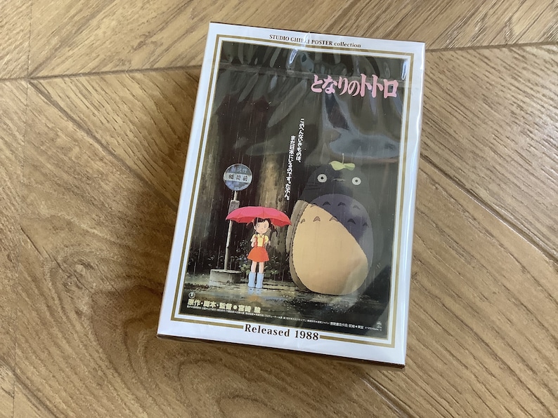 May include: A Studio Ghibli poster collection featuring the film "My Neighbor Totoro." The poster shows a young girl with a red umbrella at a bus stop, with Totoro standing nearby. The poster is in a white frame and is labeled "Released 1988."