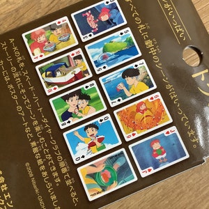 May include: A brown cardboard packaging for a deck of playing cards featuring various colorful scenes from the animated film Ponyo. The cards display characters and landscapes, with Japanese text printed on the surface and a Studio Ghibli logo.