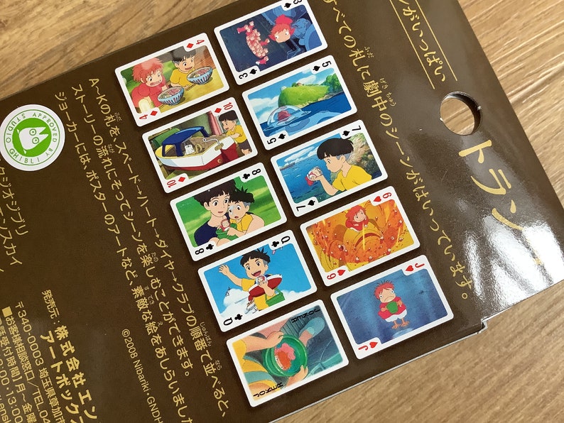 May include: A brown cardboard packaging for a deck of playing cards featuring various colorful scenes from the animated film Ponyo. The cards display characters and landscapes, with Japanese text printed on the surface and a Studio Ghibli logo.