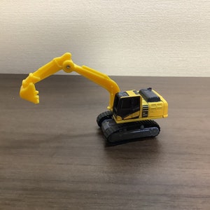 May include: A yellow and black toy excavator with a movable arm and bucket. The toy has black treads and a detailed cabin. The excavator is sitting on a brown surface.