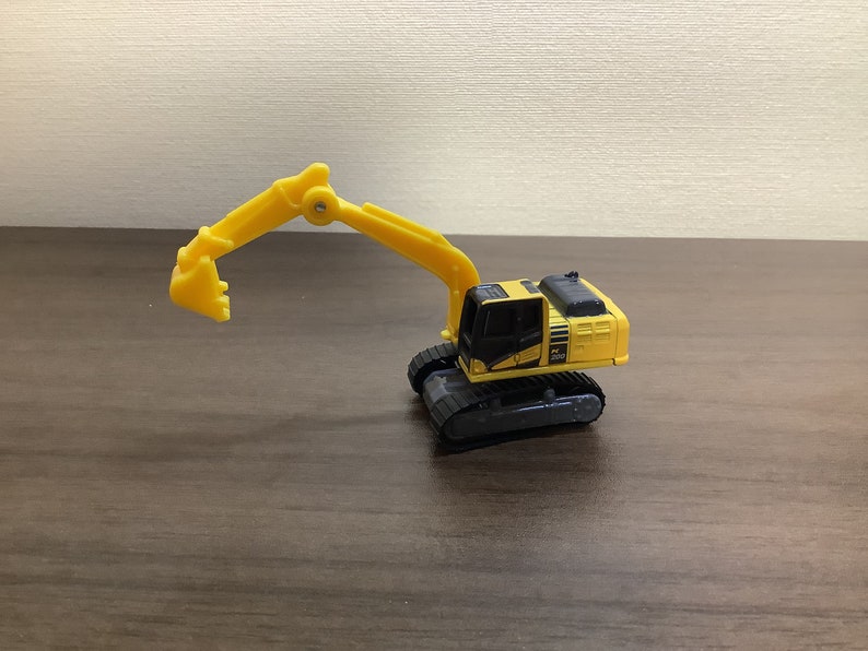 May include: A yellow and black toy excavator with a movable arm and bucket. The toy has black treads and a detailed cabin. The excavator is sitting on a brown surface.