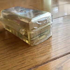 May include: A clear, rectangular plastic case with a yellow tint, likely for storing small items. The case is sealed in clear plastic wrap and sits on a wooden surface. The lid of the case has a small image on it.