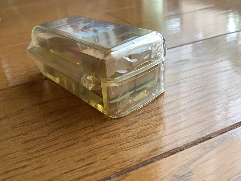 May include: A clear, rectangular plastic case with a yellow tint, likely for storing small items. The case is sealed in clear plastic wrap and sits on a wooden surface. The lid of the case has a small image on it.