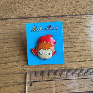 May include: A metal enamel pin featuring a cartoon character with orange hair and a red hood, mounted on a blue backing card with the Japanese text 崖の上のポニョ and English text Ponyo on the Cliff by the Sea, next to a ruler showing 5 cm.
