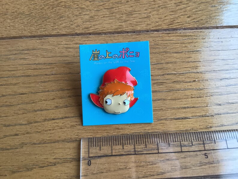 May include: A metal enamel pin featuring a cartoon character with orange hair and a red hood, mounted on a blue backing card with the Japanese text 崖の上のポニョ and English text Ponyo on the Cliff by the Sea, next to a ruler showing 5 cm.