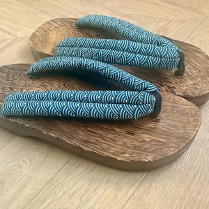May include: A pair of traditional Japanese geta sandals. The wooden platforms are a rich brown, and the straps are teal with a white wave pattern. The sandals are designed for outdoor wear.