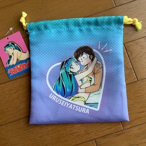 Könnte beinhalten: Ein Kordelzugbeutel mit einem blau-violetten Farbverlauf, der ein Cartoon-Paar zeigt, das sich in einem Herzen umarmt. Die Tasche hat einen gelben Kordelzug und ein Etikett mit einer Charakterillustration.