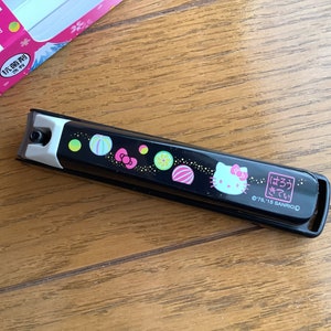 Może przedstawiać: Czarne obcinaczki do paznokci z wzorem Hello Kitty. Obcinaczki mają różową twarz Hello Kitty, różową kokardkę i kolorowe wzory japońskich cukierków. Tekst "はろうきてい" i "0176, 15 SANRIO" jest nadrukowany na obcinaczkach.