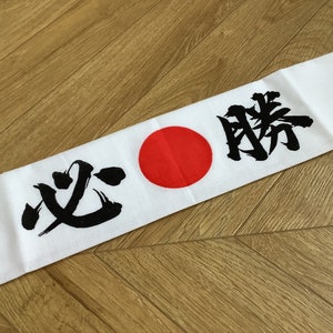 May include: White headband featuring Japanese calligraphy and a red circle. The black characters translate to "certain victory". The headband is made of white fabric.
