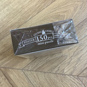 May include: A Studio Ghibli mini puzzle collection box, wrapped in clear plastic. The box is brown with white text that reads "POSTER 150 pcs. mini puzzle" and the Studio Ghibli logo. The box is on a wooden surface.