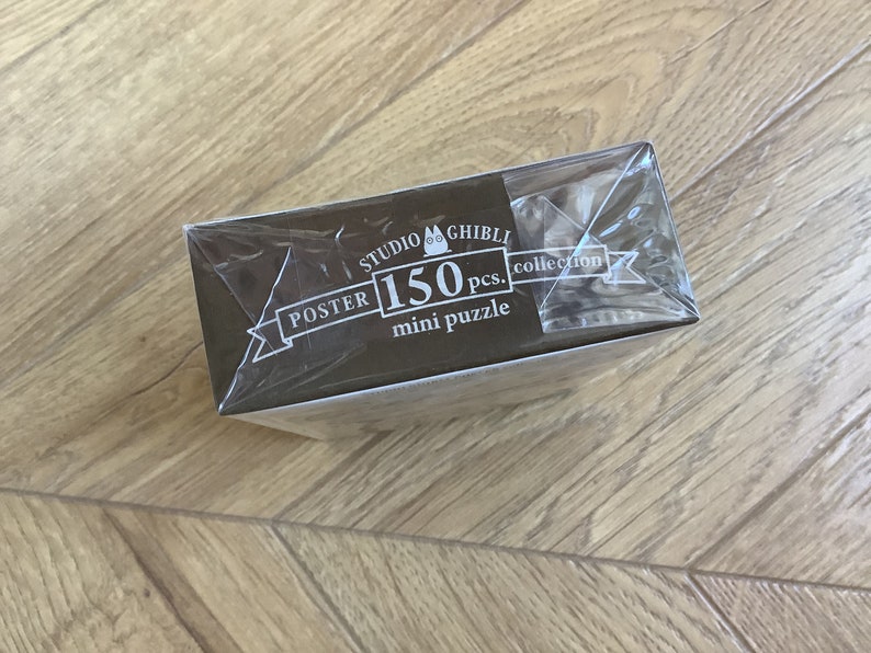 May include: A Studio Ghibli mini puzzle collection box, wrapped in clear plastic. The box is brown with white text that reads "POSTER 150 pcs. mini puzzle" and the Studio Ghibli logo. The box is on a wooden surface.