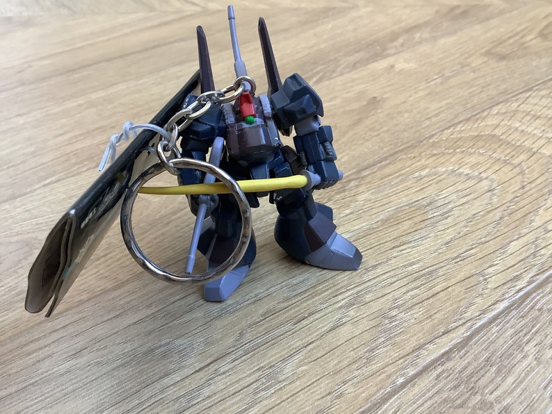 May include: A dark blue and gray robot figure keychain with a silver key ring. The robot has a detailed design with multiple antennas and a yellow beam saber. A tag is attached to the keychain.