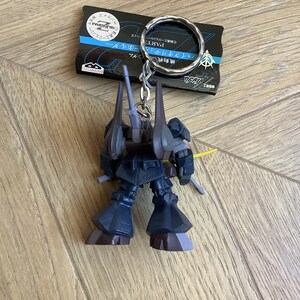 May include: A dark blue Gundam robot keychain with a silver keyring and a black tag. The robot has a yellow sword and gray accents. The tag has Japanese text and a logo.