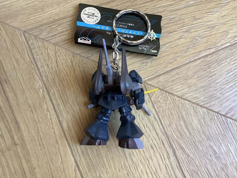 May include: A dark blue Gundam robot keychain with a silver keyring and a black tag. The robot has a yellow sword and gray accents. The tag has Japanese text and a logo.