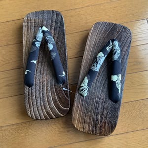 May include: A pair of geta sandals featuring dark brown, textured wooden platforms with a distinct grain pattern. The thong-style straps are made of black fabric decorated with a white and light green graphic pattern of stylized animal figures.
