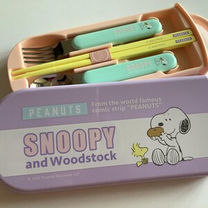 May include: A Peanuts-themed lunch set featuring Snoopy and Woodstock. The set includes a light purple case with the words "Snoopy and Woodstock" and a cartoon illustration. Inside is a peach-colored container with utensils and chopsticks.