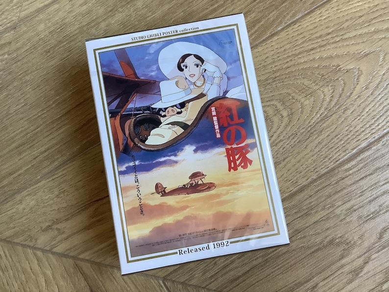 May include: A Studio Ghibli poster collection box featuring the movie "Porco Rosso." The artwork depicts a pig in a plane with a woman in a white hat. The poster has Japanese text and the release year 1992.