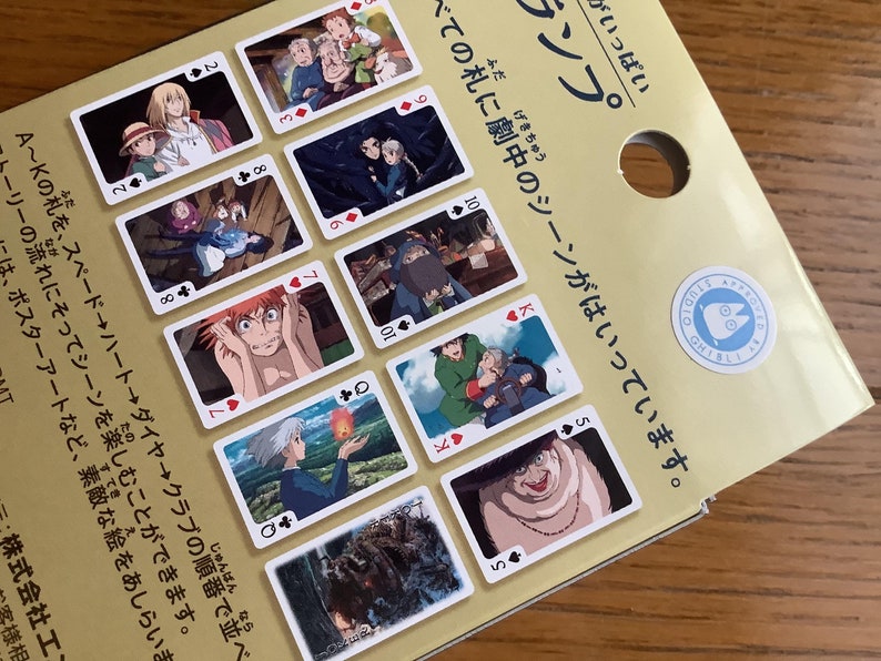 Pu&ograve; includere: Un mazzo di carte da gioco con scene di un film d'animazione giapponese. Ogni carta mostra un'immagine diversa del film, con semi e numeri di carte tradizionali. Le carte sono su uno sfondo marrone chiaro.