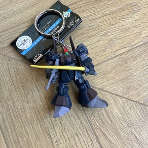 May include: A dark blue and brown robot keychain with a yellow sword. The keychain has a silver ring and a tag with Japanese text. The robot has a detailed design and is posed with its sword.
