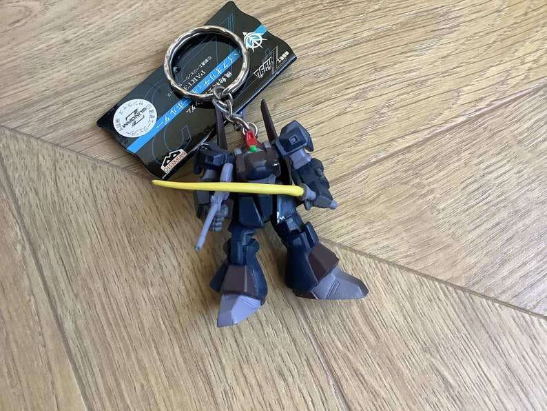 May include: A dark blue and brown robot keychain with a yellow sword. The keychain has a silver ring and a tag with Japanese text. The robot has a detailed design and is posed with its sword.