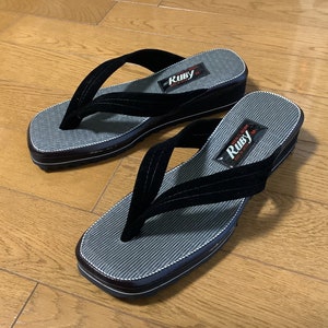 Könnte beinhalten: Ein Paar schwarze Flip-Flops mit einem grau-weiß gestreiften Fußbett. Die Flip-Flops haben einen schwarzen Riemen und ein kleines Etikett mit der Aufschrift "Ruby". Die Sohlen sind dunkelbraun mit weiß-blauer Umrandung.