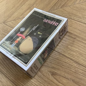 May include: A boxed Studio Ghibli poster collection featuring the animated film "My Neighbor Totoro." The box art depicts a scene with Totoro, a girl with an umbrella, and a bus stop. Released in 1988. The box size is 10 x 14.2 cm.
