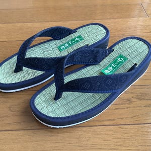 Könnte beinhalten: Ein Paar japanische Geta-Sandalen. Die Sandalen haben ein hellgrünes, gewebtes Fußbett, einen marineblauen Riemen mit Blumenmuster und eine marineblaue und weiße Sohle. Die Sandalen liegen auf einer Holzoberfläche.