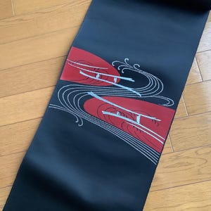 Può includere: Un tessuto nero con un design rosso e bianco. Il design presenta due forme rosse con linee bianche e un motivo a onde. Il tessuto è appoggiato su una superficie di legno.