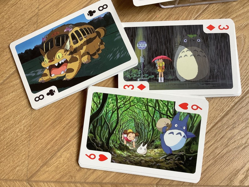 May include: Playing cards featuring scenes from the animated film, My Neighbor Totoro. The cards show images of Totoro, the Catbus, and other characters in various scenes. The cards are on a wooden surface.