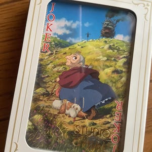 Pu&ograve; includere: Carta da gioco Joker con un'illustrazione de Il castello errante di Howl dello Studio Ghibli. La carta mostra una donna anziana con un cane, un bastone e il castello sullo sfondo. La parola 'JOKER' &egrave; stampata.
