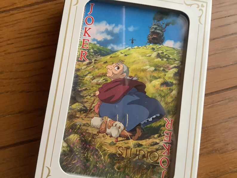 Pu&ograve; includere: Carta da gioco Joker con un'illustrazione de Il castello errante di Howl dello Studio Ghibli. La carta mostra una donna anziana con un cane, un bastone e il castello sullo sfondo. La parola 'JOKER' &egrave; stampata.