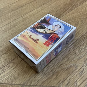 May include: A rectangular box featuring a colorful illustration from the animated film "Porco Rosso." The artwork depicts a woman in a white hat and a biplane flying in a sunset sky. The text "Released 1992" is visible.