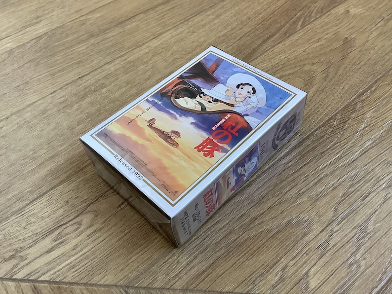 May include: A rectangular box featuring a colorful illustration from the animated film "Porco Rosso." The artwork depicts a woman in a white hat and a biplane flying in a sunset sky. The text "Released 1992" is visible.