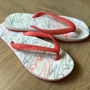 Könnte beinhalten: Weisse Flip-Flops mit roten Riemen und einem Muster aus grauen Krokodilen. Der Markenname "Lacoste" ist auf dem Riemen zu sehen. Die Sohle ist weiss mit rotem Rand. Ein Paar Sommerschuhe.