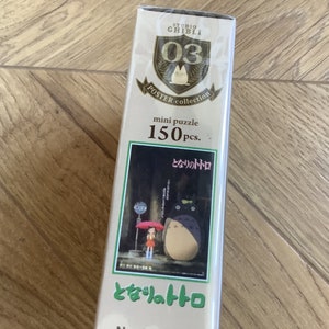 May include: A mini puzzle box from the Studio Ghibli Poster Collection featuring a scene from the movie "My Neighbor Totoro." The box is white with gold accents and contains 150 pieces. The image on the box shows Totoro and a girl under an umbrella.