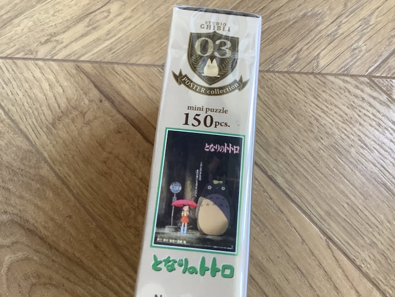 May include: A mini puzzle box from the Studio Ghibli Poster Collection featuring a scene from the movie "My Neighbor Totoro." The box is white with gold accents and contains 150 pieces. The image on the box shows Totoro and a girl under an umbrella.