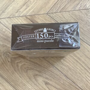 May include: A brown box for a 150-piece mini puzzle from the Studio Ghibli collection. The box is wrapped in clear plastic and features white text that reads "POSTER" and "mini puzzle".