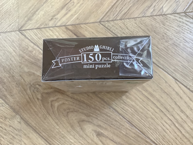 May include: A brown box for a 150-piece mini puzzle from the Studio Ghibli collection. The box is wrapped in clear plastic and features white text that reads "POSTER" and "mini puzzle".