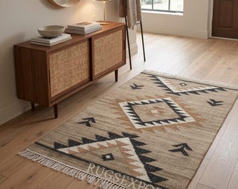 May include: A rectangular area rug featuring a woven tan base with a geometric pattern in black, white, and brown. The design includes diamond and chevron motifs with a fringed edge on the short sides. The text RUGSEXPORTS is visible.