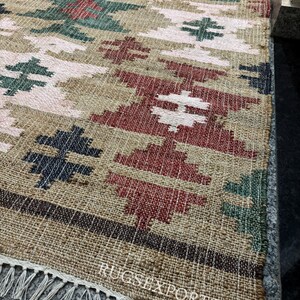 Puede incluir: Una alfombra tejida con un patr&oacute;n geom&eacute;trico en tonos beige, rojo, verde y azul marino. La alfombra tiene flecos en un borde y el texto "RUGSEXPORTS" es visible. La alfombra est&aacute; hecha de fibra natural.