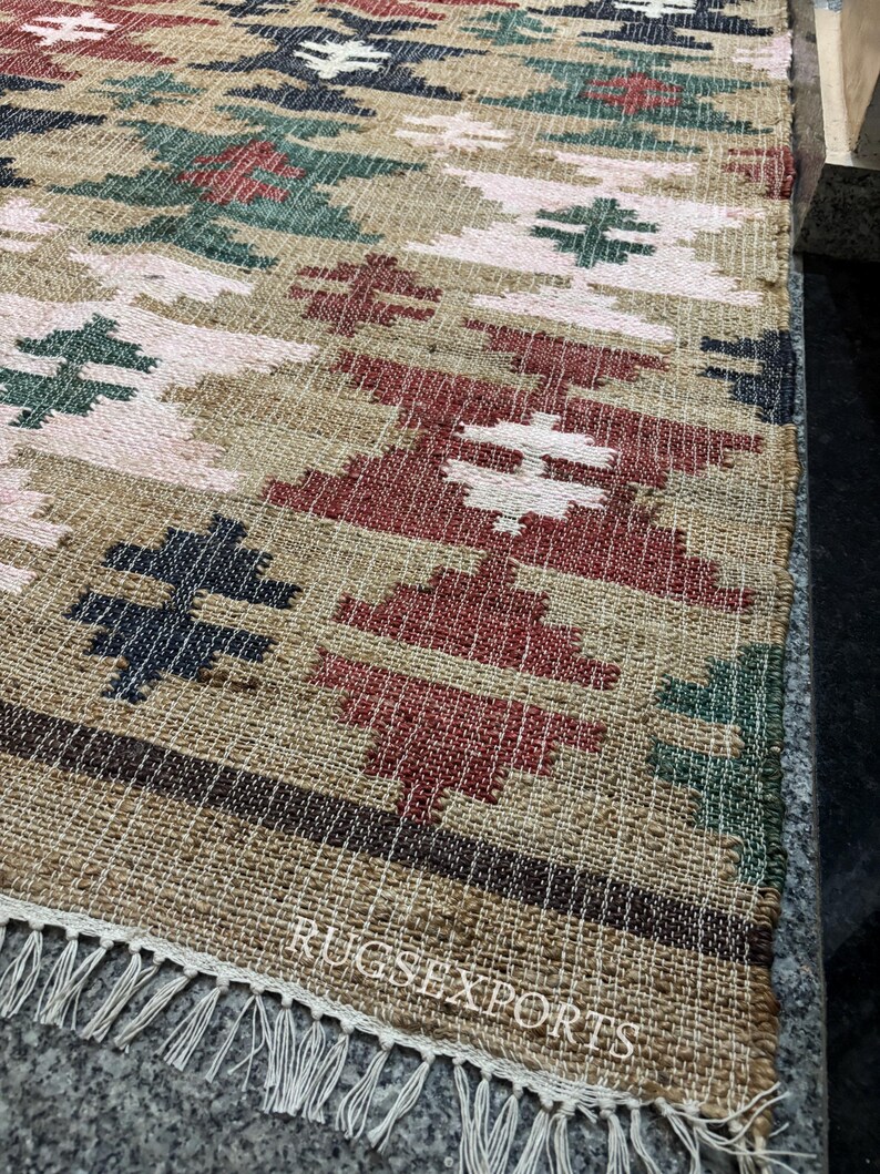 Puede incluir: Una alfombra tejida con un patr&oacute;n geom&eacute;trico en tonos beige, rojo, verde y azul marino. La alfombra tiene flecos en un borde y el texto "RUGSEXPORTS" es visible. La alfombra est&aacute; hecha de fibra natural.