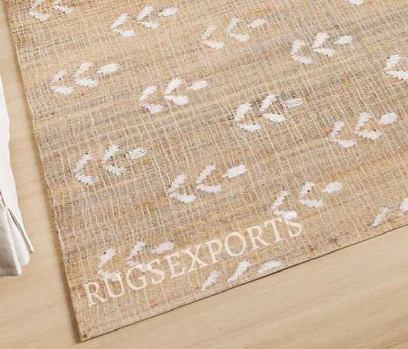 May include: A beige and white area rug with a woven texture and a white arrow pattern. The rug has the text "RUGSEXPORTS" printed on it.